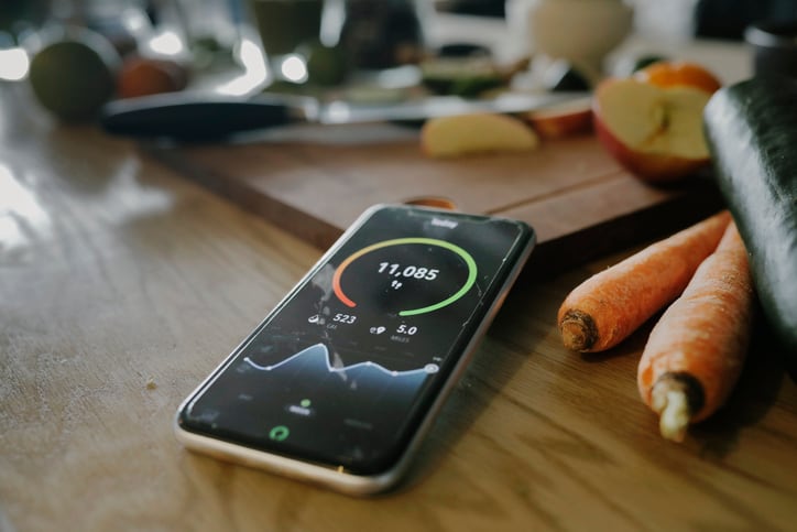 A close up of a smartphone laying on a chopping board next some vegetables while displaying a health app on screen.