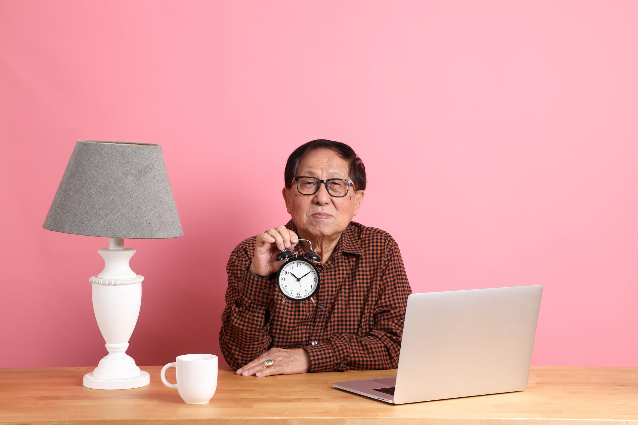 A senior man sitting in front of his work desk and holding a clock.