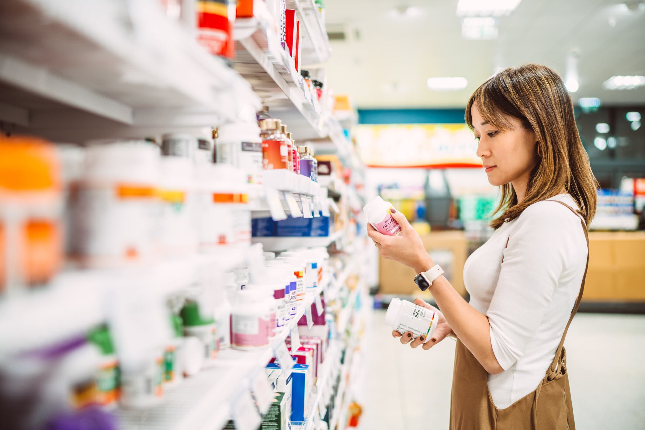 Asian woman examining bottles of vitamins and dietary supplements in a pharmacy or supermarket aisle. She is comparing labels and making a decision on health and wellness products. Concept of healthy lifestyle, nutrition, self-care, and wellness.